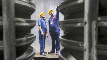 Two male Sodexo employees in overalls and hard hats pointing up to the top of machinery