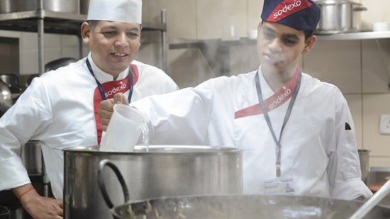 Two sodexo chefs pouring liquid in to a saucepan