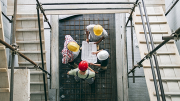 Four adults in a construction site wearing hard hats and looking at building plans, photographed from above